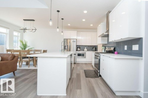Open-concept kitchen featuring an island with a white countertop, white cabinetry with polished hardware, stainless steel appliances, a grey subway tile backsplash, and wood-finish flooring - 5627 Kootook Place, Edmonton, AB - Indoor Photo Showing Kitchen With Upgraded Kitchen