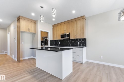 Kitchen featuring a central island with a dark countertop and undermount sink, light wood-finish cabinetry, stainless steel microwave, black hexagonal tile backsplash, and recessed lighting - 3714 40 Avenue, Beaumont, AB - Indoor Photo Showing Kitchen With Upgraded Kitchen