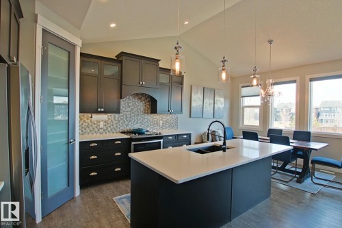 Open-concept kitchen featuring dark cabinetry, a white countertop island with an undermount sink, a mosaic tile backsplash, and stainless steel appliances - 9711 223 Street, Edmonton, AB 