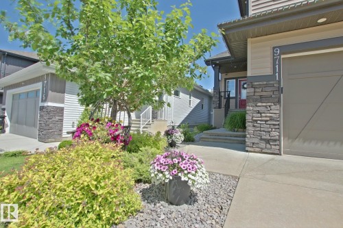 Front exterior featuring a stone-clad garage and entryway pillars, a red entry door, a paved walkway, and landscaping with mature trees and flowering shrubs - 9711 223 Street, Edmonton, AB 