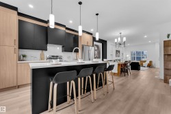 Open-concept kitchen featuring a spacious island with seating, two-tone cabinetry with light wood-finish and matte black panels, stainless steel appliances, and wood-finish flooring - 