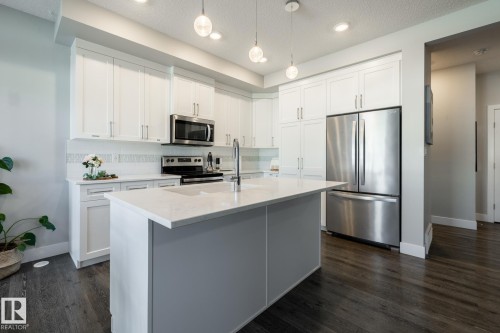 Contemporary kitchen featuring white shaker cabinetry, stainless steel appliances, a center island with a light-toned countertop, a geometric tile backsplash, and dark wood-finish flooring - 1440 Darby Green, Edmonton, AB - Indoor Photo Showing Kitchen With Upgraded Kitchen
