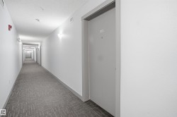 Corridor featuring patterned carpet flooring, white painted walls, wall-mounted light fixtures, and a gray door with a visible unit number - 