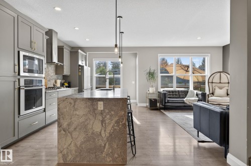 Open-concept kitchen and living area featuring a large central island with stone countertop, gray cabinetry, stainless steel appliances including a built-in oven and microwave, and wood-finish flooring - 1737 Dumont Crescent, Edmonton, AB - Indoor Photo Showing Kitchen With Upgraded Kitchen