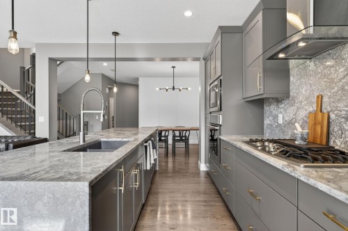 Spacious kitchen featuring gray cabinetry with gold-tone hardware, a large island with an undermount sink, stone countertops and backsplash, and a built-in gas range with a modern range hood - 1737 Dumont Crescent, Edmonton, AB - Indoor Photo Showing Kitchen With Upgraded Kitchen