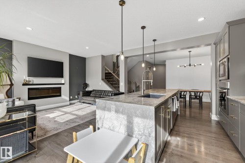 Open-concept living area featuring a large kitchen island with integrated sink, wood-finish flooring, a contemporary fireplace with a floating shelf, and a staircase with dark wood treads and risers - 1737 Dumont Crescent, Edmonton, AB - Indoor Photo Showing Kitchen With Upgraded Kitchen