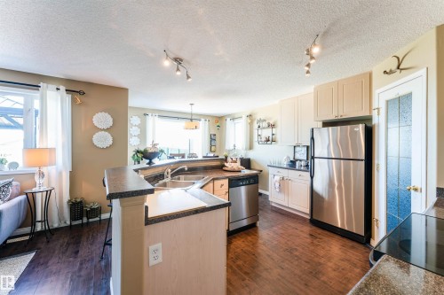 Open-concept kitchen featuring wood-finish flooring, light-toned cabinetry, stainless steel appliances, track lighting, and a breakfast bar with dark countertops - 847 Blacklock Way, Edmonton, AB - Indoor Photo Showing Kitchen With Double Sink