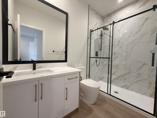 Modern bathroom featuring a vanity with a white countertop and gold hardware, a black-framed mirror, and a walk-in shower with a glass enclosure and marble-patterned walls - 17 Trill Point(E), Spruce Grove, AB - Indoor Photo Showing Bathroom