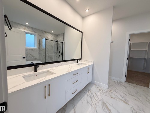 The bathroom features a large vanity with dual sinks, white cabinetry, and gold-toned hardware, all beneath a generously sized mirror with a black frame - 17 Trill Point(E), Spruce Grove, AB - Indoor Photo Showing Bathroom