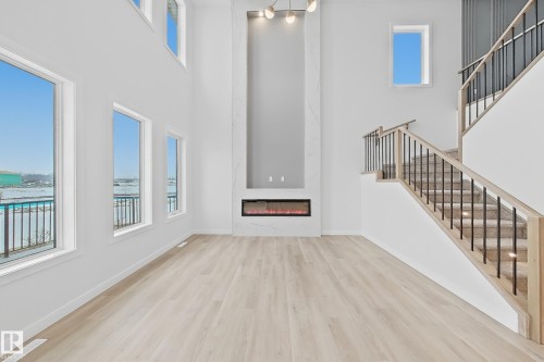 This spacious living area features light-toned flooring, a contemporary fireplace with an integrated entertainment wall, and a staircase with light wood handrails and black metal balusters - 17 Trill Point(E), Spruce Grove, AB - Indoor Photo Showing Other Room