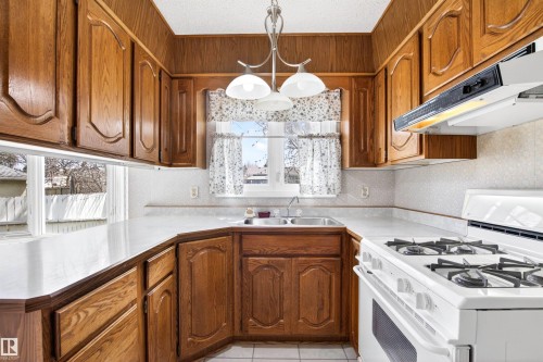 U-shaped kitchen featuring wood cabinetry, white laminate countertops, a double basin sink, white gas range with integrated exhaust hood, and white tile flooring - 29 Bristow Crescent, Spruce Grove, AB - Indoor Photo Showing Kitchen With Double Sink