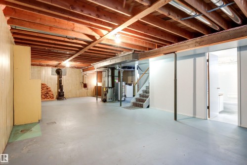 Expansive basement space featuring exposed wood joists, painted concrete flooring, vertical wood paneling, and a corner wood-burning stove with brick storage - 29 Bristow Crescent, Spruce Grove, AB - Indoor Photo Showing Basement