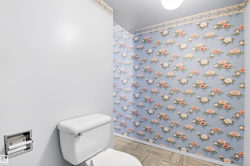 Powder room featuring a white toilet, recessed toilet paper holder, patterned wallpaper with floral motif, and wood-finish flooring - 29 Bristow Crescent, Spruce Grove, AB - Indoor Photo Showing Bathroom