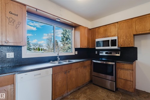 Kitchen featuring ample wood-finish cabinetry, a large window, a double basin stainless steel sink, black countertops, and a white dishwasher - 3752 114 Street, Edmonton, AB - Indoor Photo Showing Kitchen With Double Sink