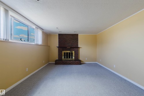 Spacious interior featuring a brick fireplace with a brass-trimmed firebox, light beige walls, a neutral grey carpet, and a large window with white blinds - 3752 114 Street, Edmonton, AB - Indoor With Fireplace