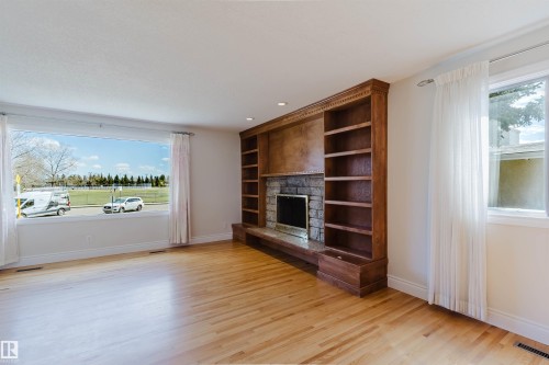 Living area featuring wood-finish flooring, a broad picture window, and a built-in entertainment center with a stone-surround fireplace and integrated shelving - 3752 114 Street, Edmonton, AB - Indoor Photo Showing Living Room With Fireplace
