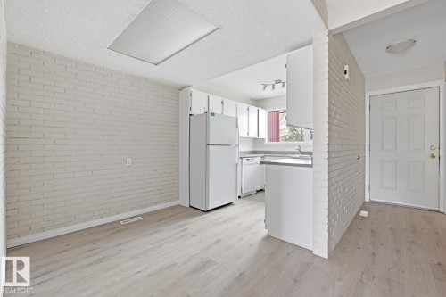 Open-concept living space featuring wood-finish flooring, white brick-finish accent walls, a kitchen with white cabinetry, a white refrigerator, and a front entry door with paneling - 10640 Beaumaris Road, Edmonton, AB - Indoor Photo Showing Kitchen