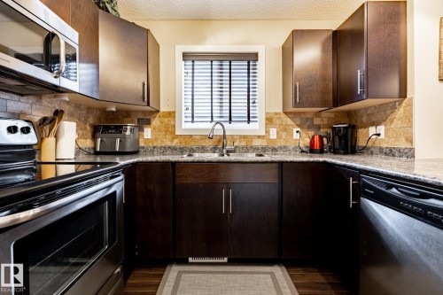 The property features a kitchen with dark wood cabinetry, a tiled backsplash, and stainless steel appliances including a microwave, oven, and dishwasher - 40 12004 22 Avenue, Edmonton, AB - Indoor Photo Showing Kitchen With Double Sink