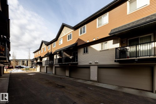 The property features a two-story exterior with brown and beige siding, a private balcony with black railings, and an attached garage with a brown door - 40 12004 22 Avenue, Edmonton, AB - Outdoor