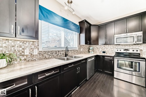 Kitchen featuring dark wood-finish cabinetry, stainless steel appliances, mosaic tile backsplash, wood-finish flooring, and a double basin sink with a matte black faucet - 5020 Orchards Gate, Edmonton, AB - Indoor Photo Showing Kitchen With Stainless Steel Kitchen With Double Sink With Upgraded Kitchen
