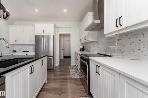Well-appointed kitchen featuring white shaker-style cabinetry with black hardware, wood-finish flooring, a stainless steel refrigerator, and dark countertops on the island - 1450 Darby Green, Edmonton, AB - Indoor Photo Showing Kitchen With Stainless Steel Kitchen With Upgraded Kitchen