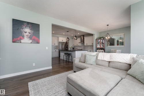 Open-concept living area featuring wood-finish flooring, a kitchen with white cabinetry, a kitchen island with seating, pendant lighting, and a dining area with a geometric chandelier - 1450 Darby Green, Edmonton, AB - Indoor Photo Showing Living Room