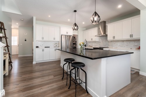Contemporary kitchen featuring a large central island with a dark countertop, white perimeter cabinetry with dark hardware, a stainless steel range hood, wood-finish flooring, and recessed lighting - 1450 Darby Green, Edmonton, AB - Indoor Photo Showing Kitchen With Stainless Steel Kitchen With Upgraded Kitchen