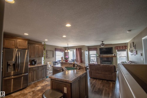 Open-concept living space featuring a kitchen with wood cabinetry and a stainless-steel refrigerator, a central island with a sink, wood-finish flooring, and a living area with a fireplace - 5121 52 Ave, St. Paul Town, AB - Indoor Photo Showing Kitchen