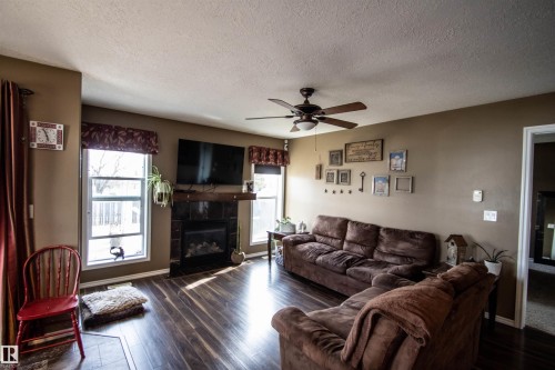 Living area featuring wood-finish flooring, a ceiling fan with integrated lighting, and a fireplace with a dark tile surround and wood mantel - 5121 52 Ave, St. Paul Town, AB - Indoor Photo Showing Living Room With Fireplace