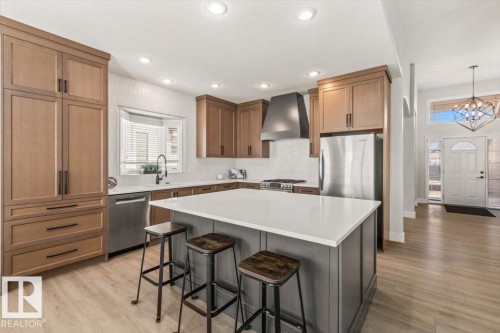 Modern kitchen featuring wood-finish cabinetry, a large center island with a white countertop, stainless steel appliances, a bay window, and wood-finish flooring - 7719 166 Avenue, Edmonton, AB - Indoor Photo Showing Kitchen With Upgraded Kitchen