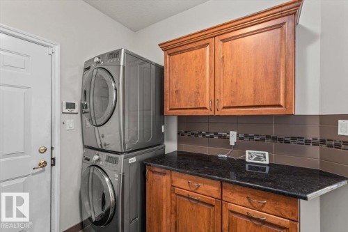 Laundry area featuring stacked front-load machines, wood-finish cabinetry, a dark countertop, and a tiled backsplash with mosaic accents - 7719 166 Avenue, Edmonton, AB - Indoor Photo Showing Laundry Room