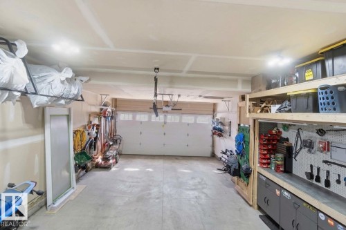 Attached two-car garage featuring a garage door with window inserts, multiple overhead lighting fixtures, a built-in workbench with cabinetry, and exposed ceiling joists - 1825 Westerra Loop, Stony Plain, AB - Indoor Photo Showing Garage