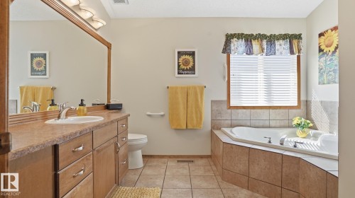 Bathroom featuring a built-in jetted tub with tiled surround, a window with blinds, a vanity with a single sink and cabinet storage, a large framed mirror, and tile flooring - 5 Richelieu Place, Beaumont, AB - Indoor Photo Showing Bathroom