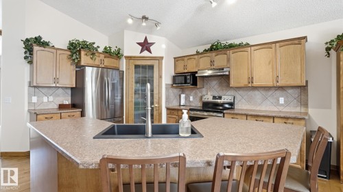 Kitchen featuring an angled island with an undermount sink and a gooseneck faucet, light wood-finish cabinetry, a stainless steel refrigerator, and a tile backsplash - 5 Richelieu Place, Beaumont, AB - Indoor Photo Showing Kitchen