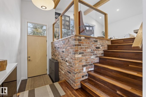 Entryway featuring a light-colored door with an overhead transom window, a prominent exposed brick wall, and rich wood stairs with a matching wood railing - 9736 78 Avenue, Edmonton, AB - Indoor Photo Showing Other Room