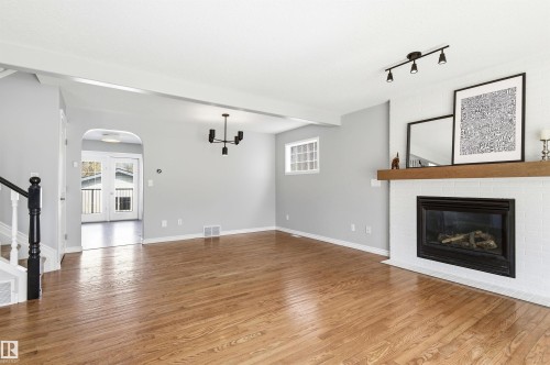 Spacious living area featuring polished wood-finish flooring, a white tiled fireplace with a wood-beam mantel, and recessed window - 8730 92A Avenue, Edmonton, AB - Indoor Photo Showing Living Room With Fireplace