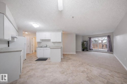 Open-concept living space featuring a kitchen with white cabinetry and light-toned countertops, tile flooring, and a carpeted living area with a sliding glass door providing exterior access - 103 12110 119 Ave, Edmonton, AB - Indoor Photo Showing Kitchen