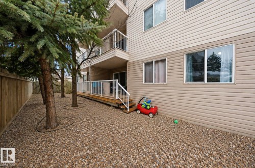 Ground-level patio with wood decking and white metal railings, set against a gravel landscape featuring mature evergreen trees - 103 12110 119 Ave, Edmonton, AB - Outdoor With Balcony With Exterior