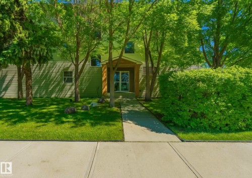 Entryway featuring a wood-clad portico with double glass doors, surrounded by mature trees, a manicured lawn, and lush green shrubbery - 103 12110 119 Ave, Edmonton, AB - Outdoor