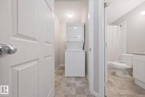 Dedicated laundry area with stacked washer and dryer, adjacent to a bathroom featuring a white subway tile shower surround and a vanity with a light-toned countertop - 103 12110 119 Ave, Edmonton, AB - Indoor Photo Showing Other Room