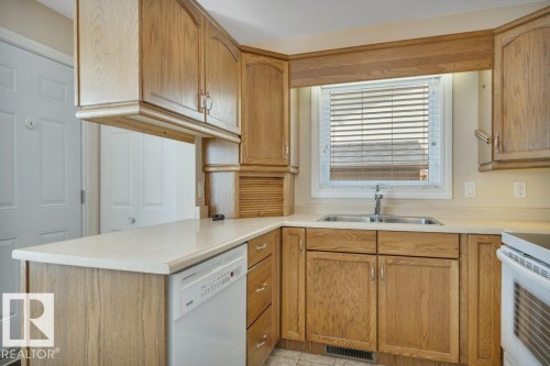 Kitchen featuring wood-finish cabinetry, light-toned countertops, and a double-basin stainless steel sink under a window with blinds - 18227 76 Avenue, Edmonton, AB - Indoor Photo Showing Kitchen With Double Sink