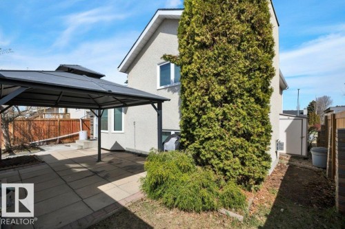 Paved patio area featuring a dark-framed gazebo, exterior stucco finish, mature evergreen shrubbery, and a compact storage shed - 18227 76 Avenue, Edmonton, AB - Outdoor