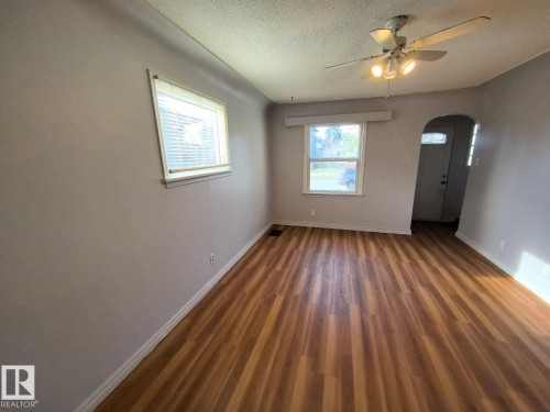 Spacious room featuring wood-finish flooring, two windows with blinds, a ceiling fan with integrated lighting, and an arched entryway - 10815 68 Avenue, Edmonton, AB - Indoor Photo Showing Other Room