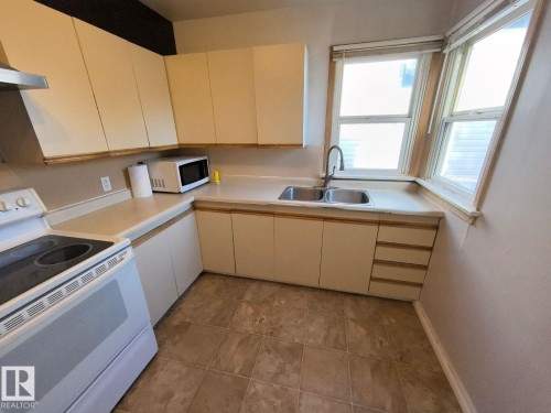 Kitchen featuring a dual-basin stainless steel sink, a corner window configuration, cream cabinetry, light-toned countertops, and tiled flooring - 10815 68 Avenue, Edmonton, AB - Indoor Photo Showing Kitchen With Double Sink