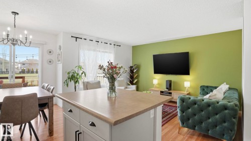 Open-concept living area featuring wood-finish flooring, a kitchen island with a light-toned countertop, a dining area with chandelier lighting, a large window with sheer drapery, and a sliding glass door providing exterior access - 80 287 Macewan Road, Edmonton, AB - Indoor