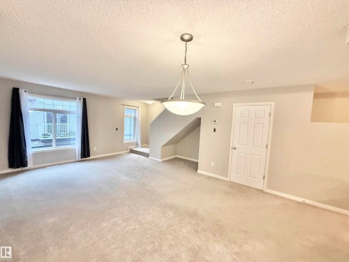 Spacious carpeted room featuring a large window, a modern pendant light fixture, a staircase with carpeted treads, and a paneled interior door - 97 465 Hemingway Road, Edmonton, AB - Indoor Photo Showing Other Room