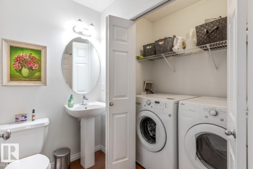 Convenient laundry closet with stacked appliances and wire shelving, complemented by a pedestal sink, oval mirror, and wall-mounted light fixture - 7641 Schmid Crescent, Edmonton, AB - Indoor Photo Showing Laundry Room