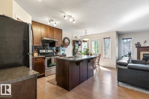 Open-concept living area featuring wood-finish flooring, a kitchen island with integrated sink, stainless steel appliances, and a built-in fireplace with wood mantel - 7641 Schmid Crescent, Edmonton, AB - Indoor Photo Showing Kitchen With Fireplace