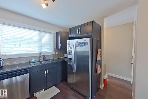 Kitchen featuring dark wood-finish cabinetry, stainless steel appliances, dark countertop, dual basin sink, and a tiled backsplash - 295 401 Southfork, Leduc, AB - Indoor Photo Showing Kitchen With Double Sink