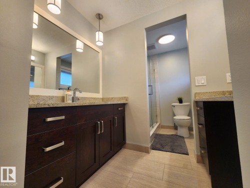 Bathroom featuring dark wood-finish cabinetry with polished chrome hardware, light-toned granite countertops, a large framed mirror with integrated lighting, and a glass-enclosed shower - 12423 28A Avenue, Edmonton, AB - Indoor Photo Showing Bathroom